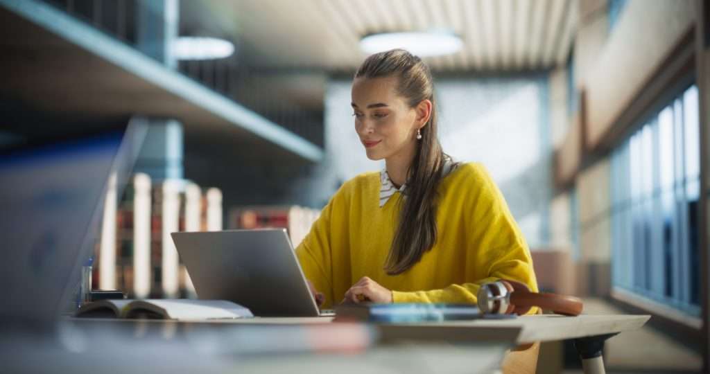 Woman sat in library environment working on a laptop on the table in front of her. She is fressed in a yellow jumper and there is a n open book and headphones by her. The background is blurred with the focus on the woman.