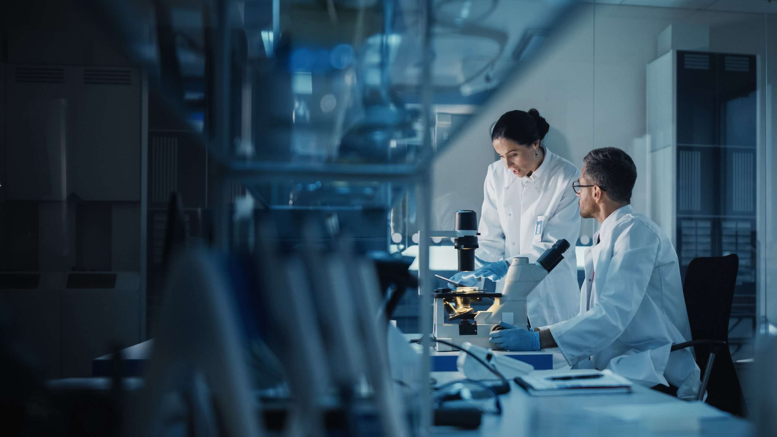 Modern Medical Research Laboratory: Two Scientists Working, Using Digital Tablet, Analyzing Test, Talking. Advanced Scientific Pharmaceutical Lab for Medicine, Biotechnology Development. Evening Time